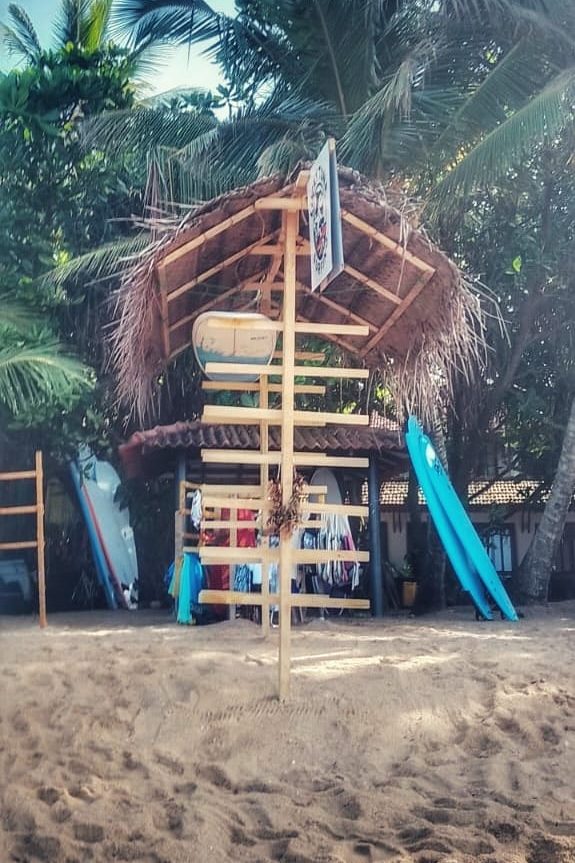 Ein surfender Holzstand mit einem Strohdach, umgeben von Palmen und Sand, im Hintergrund sind Surfbretter zu sehen.