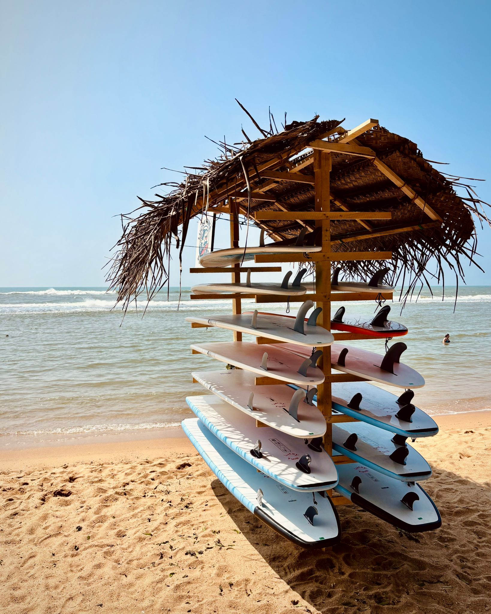 Ein Surfbrettständer mit mehreren Surfboards am Strand, im Hintergrund das Meer und ein klarer blauer Himmel.